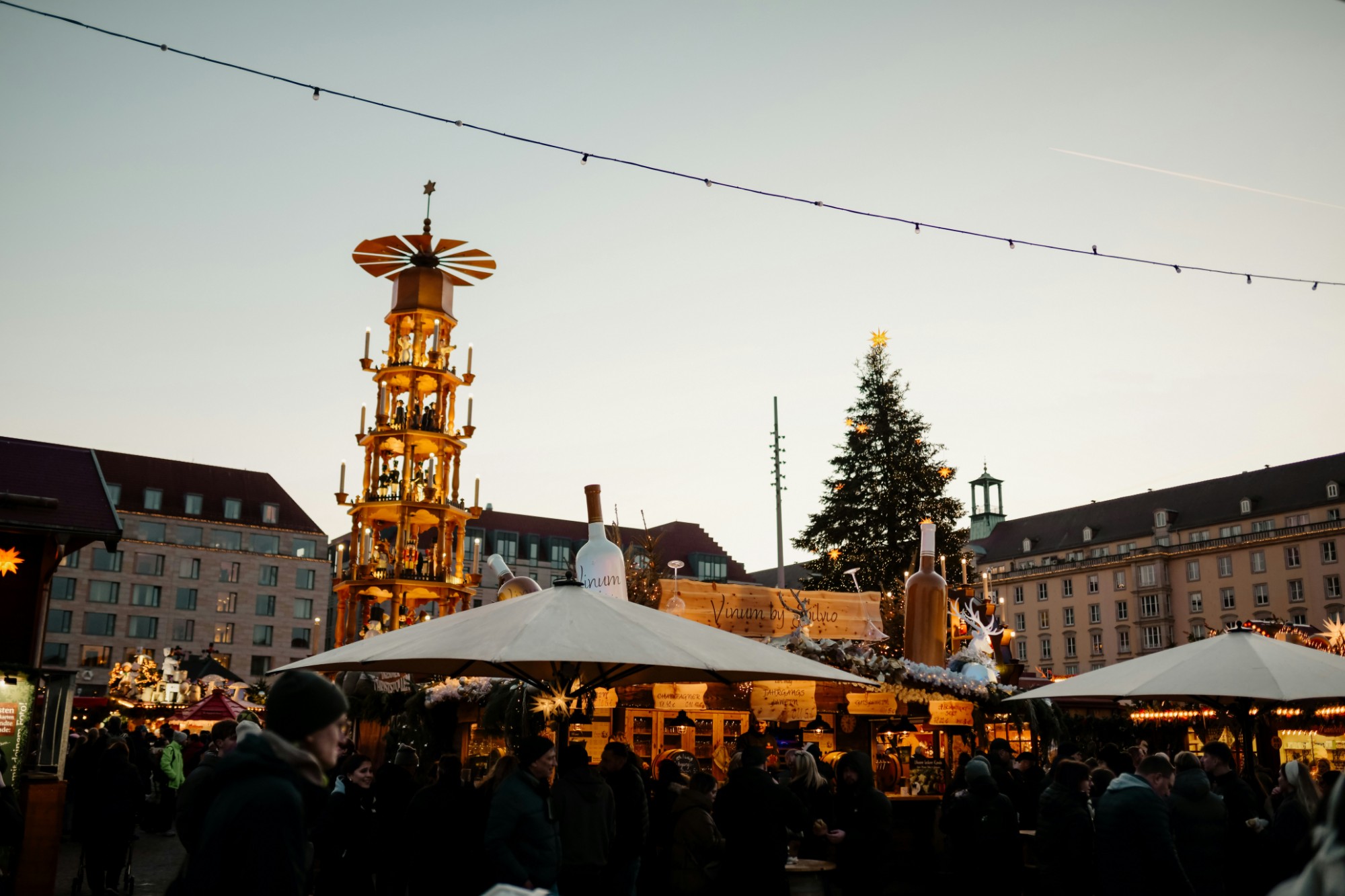 A Christmas market at dusk, with a lit wooden Christmas pyramid, a decorated tree and crowded stalls